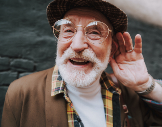 A man with a white beard smiling as he holds a cupped hand to his ear.