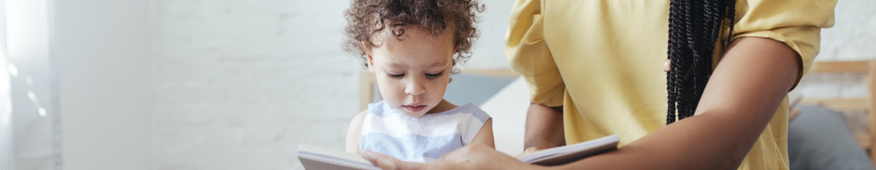 A young girl looks at a book that her mother holds open for her.